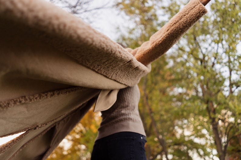 May include: A person wearing a brown fuzzy coat with the sleeves spread out, standing in a forest with fall foliage.