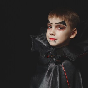 May include: A young child dressed as a vampire for Halloween. They are wearing a black cape with a red lining and have vampire makeup on their face.