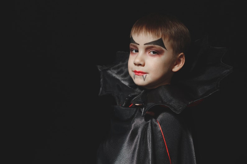 May include: A young child dressed as a vampire for Halloween. They are wearing a black cape with a red lining and have vampire makeup on their face.
