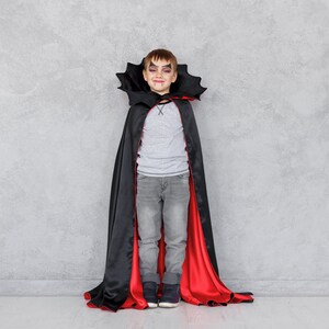 May include: A young boy dressed as a vampire for Halloween. He is wearing a black cape with a red lining and has vampire makeup on his face.