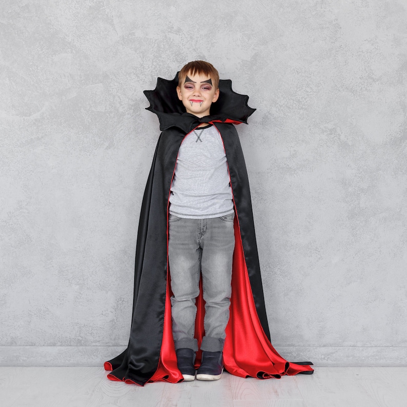 May include: A young boy dressed as a vampire for Halloween. He is wearing a black cape with a red lining and has vampire makeup on his face.