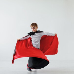 May include: A young person dressed as a vampire for Halloween. They are wearing a black and red cape with a black collar. The cape is flowing behind them as they pose for a photo.