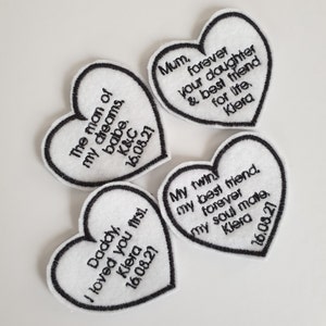 May include: Four white felt heart-shaped patches with black embroidered text. The text on the patches reads: "The man of my dreams, babe. K&C 16.08.21", "Daddy, I loved you first. Kiera 16.08.21", "Mum. Forever your daughter & best friend for life. Kiera", and "My twin my best friend. Forever my soul mate. Kiera 16.08.21".