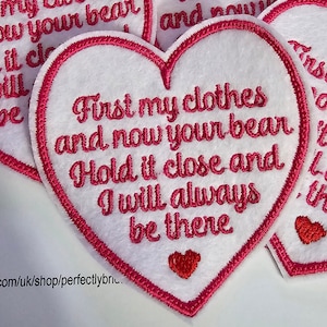 May include: A white heart-shaped patch with pink stitching and a pink embroidered message that reads "First my clothes and now your bear Hold it close and I will always be there". There is a small red heart at the bottom of the patch.