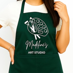 Op de afbeelding: Een donkergroene schort met een witte afbeelding van een verfpalet en penselen. De tekst "Madeline's ART STUDIO" staat in het wit. De schort, gedragen door een persoon, heeft een zak.