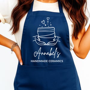 Può includere: Un grembiule blu navy con una grafica bianca di un vaso di ceramica tenuto da mani, con cuori e stelle sopra. Il grembiule ha il testo "Annabels Handmade Ceramics" in corsivo bianco.