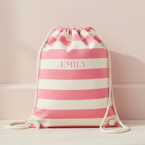 May include: A drawstring backpack featuring horizontal pink and white stripes. The center of the bag displays the name EMILY printed in a pink serif font. The bag includes thick white rope cords that cinch the top and attach at the bottom corners.