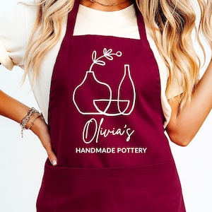 Op de afbeelding: Bordeauxrode schort met witte lijntekening van aardewerk en de tekst "Olivia's Handmade Pottery". De schort heeft een slab, een zak en lange banden.