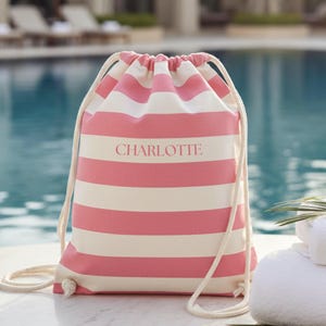 May include: A pink and white striped drawstring bag with the name "CHARLOTTE" printed on the front. The bag has a cream-colored drawstring and is sitting near a pool.