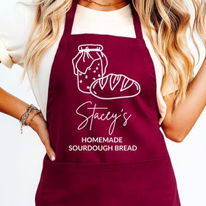 Pode incluir: Avental bordô com uma ilustração branca de um pote e um pão. O avental tem o nome "Stacey's" em escrita cursiva, com as palavras "Homemade Sourdough Bread" abaixo. O avental está sendo usado.