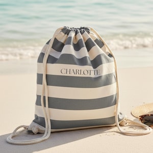 Könnte beinhalten: Grau-weiß gestreifter Turnbeutel mit dem Namen "CHARLOTTE" auf der Vorderseite. Der Beutel hat einen Kordelzugverschluss und liegt am Strand. Ideal für Strandbesuche oder zum Tragen von Gegenständen.