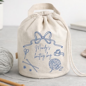 Pode incluir: Uma bolsa de tricô bege com cordão, laço azul e o texto "Mandy's knitting bag". A bolsa apresenta ilustrações azuis de ferramentas de tricô, incluindo tesouras, agulhas, uma fita métrica e uma bola de lã. Possui uma alça.