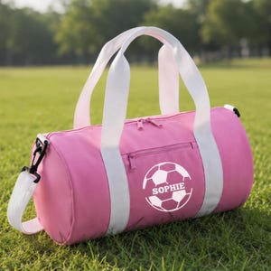Puede incluir: Una bolsa de deporte cilíndrica de color rosa con asas blancas y una correa de hombro extraíble. El lateral presenta un gráfico blanco de un balón de fútbol con el nombre SOPHIE impreso en el centro. Incluye un bolsillo lateral con cremallera.