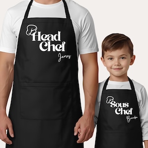 Op de afbeelding: Zwarte schorten met witte tekst. Op één schort staat "Head Chef James" en op de andere "Sous Chef Brandon". Beide schorten hebben een koksmutsymbool. De schorten hebben een grote voorzak.