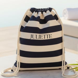 Può includere: Un sacco con coulisse a righe orizzontali blu navy e bianche. La parola "JULIETTE" è stampata in nero sul davanti. La borsa ha un cordoncino color crema. La borsa è su una superficie bianca.
