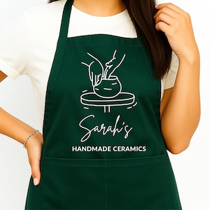 Puede incluir: Delantal verde oscuro con un gráfico blanco de manos trabajando arcilla en un torno de alfarero. El delantal tiene el texto "Sarah's Handmade Ceramics" en escritura blanca y letras de imprenta. El delantal tiene un bolsillo y una correa para el cuello.