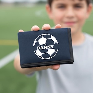 Pode incluir: Carteira azul marinho com um design de bola de futebol branca e o nome "DANNY" impresso em branco. A carteira tem uma borda preta. O fundo é um campo verde.