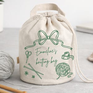 Pode incluir: Uma bolsa de tricô bege com cordão, laço verde e a frase "Emmeline's knitting bag". A bolsa apresenta ilustrações verdes de tesouras, agulhas de tricô, fita métrica, novelo de lã e flores. Um novelo de lã cinza é visível.