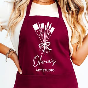 Può includere: Un grembiule bordeaux con una grafica bianca di pennelli legati con un fiocco. Il grembiule presenta il testo "Olivia's ART STUDIO" in un carattere corsivo. Il grembiule è indossato da una persona.