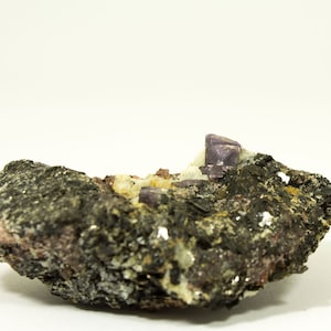 Op de afbeelding: Een close-up van een paars mineraalmonster dat is ingebed in een donkergrijze rotsmatrix. Het mineraal heeft een scherpe, kristallijne structuur en is omgeven door kleinere, witte kristallen.