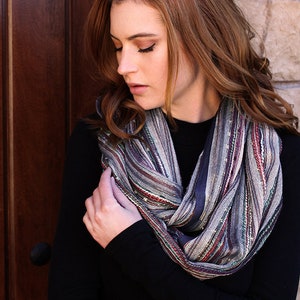 May include: A woman wearing a black turtleneck sweater with a striped infinity scarf. The scarf is made of a soft, textured fabric and features a variety of colors, including gray, blue, red, and white.
