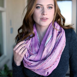 May include: A woman wearing a purple and pink striped infinity scarf. The scarf is made of a soft, lightweight fabric and has a subtle shimmer.
