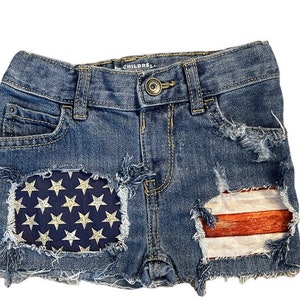 May include: Blue denim shorts with distressed details and patches featuring the American flag and stars. The shorts have a button closure, pockets, and frayed edges, perfect for a patriotic look.