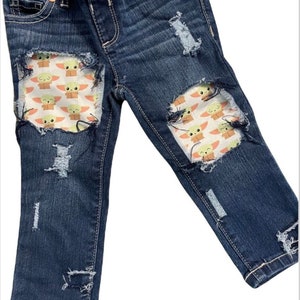 May include: Distressed blue denim jeans with patches of a cartoon character on the knees. The character is green with big eyes and a small mouth. The patches are on a white background with pink accents.