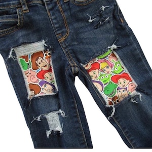 May include: Dark blue denim jeans with distressed details. Patches featuring colorful cartoon characters are sewn into the knees. The jeans have a classic five-pocket design and a button and zipper closure.