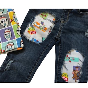 May include: Blue denim jeans with distressed patches featuring a Toy Story theme. The patches are made of white fabric with colorful Toy Story characters and the text "Toys at Play".