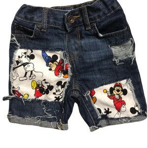 May include: Blue denim shorts with Mickey Mouse patches. The shorts have distressed details and frayed edges.