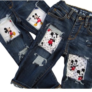 May include: Two pairs of blue denim jeans with distressed patches featuring Mickey Mouse. The patches are white with gray and red accents.