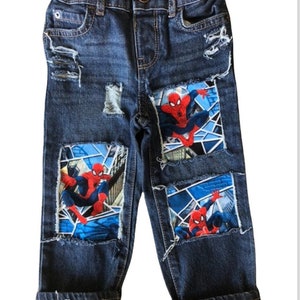 May include: Blue denim jeans with distressed patches featuring a Spiderman graphic. The patches are sewn on the front of the jeans.