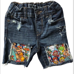 May include: Blue denim shorts with distressed details and a colorful Mario character print patch on the front.