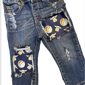 May include: Dark wash denim jeans with distressed details and patches featuring a repeating pattern of a cartoon character. The patches are a dark blue fabric with the character in tan and white.