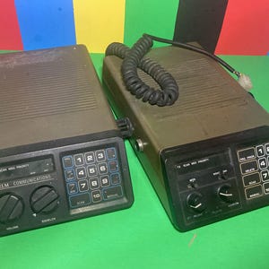 May include: Two olive-green RELM Communications radios. Each has a black control panel with a keypad, buttons, and a small display. One has a coiled cord. The radios are on a green surface.