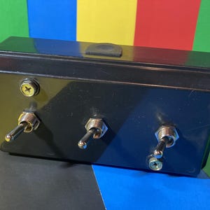 May include: A black rectangular electrical component box with three chrome toggle switches and two screws. The box is made of a glossy black material. The background is a colorful mix of green, yellow, red, blue, and black.