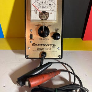 May include: Vintage Marquette Circuit Tester with a gold-colored face, black and red probes, and a black cable. The tester features a dial with voltage and ohms readings, a test selector, and the text "MARQUETTE CIRCUIT TESTER".