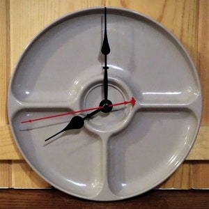 May include: A unique wall clock made from a divided serving dish. The clock face is a light gray color with black clock hands and a red second hand. The clock is mounted on a wooden surface.