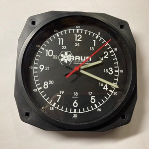 May include: Black Braun quartz clock with white numerals and hands. The clock face has a 24-hour scale and a red second hand. The clock is encased in a black, square-shaped frame with mounting holes.