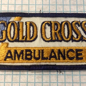 May include: Embroidered patch with the words "GOLD CROSS AMBULANCE" in gold thread on a white background. The patch has a navy blue border and a gold and blue design element.