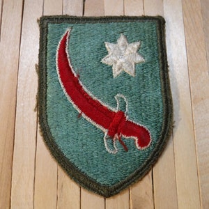 May include: A green embroidered patch with a red scimitar and a white star. The patch is shaped like a shield.