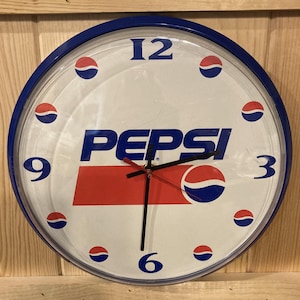 May include: Round Pepsi clock with a white face, blue bezel, and black hands. The clock features the Pepsi logo in blue and red, with the numbers 12, 3, 6, and 9 in blue. Red and blue Pepsi logo accents are around the clock face.