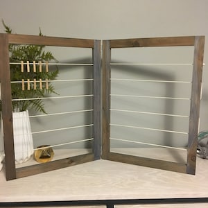 May include: A wooden picture frame with a white string grid and gold clothespins. The frame is divided into two sections and is standing on a light wood surface.
