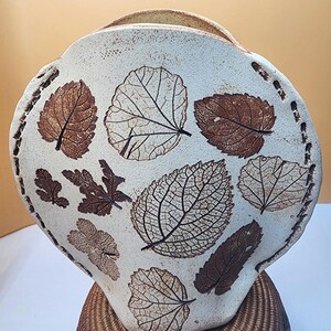 May include: A round, off-white ceramic vase featuring a flat, wide front surface decorated with various brown leaf imprints. The edges of the vase are accented with a series of small, dark brown indentations, and the top has a wide, open mouth.
