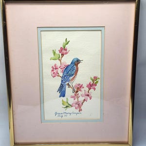 May include: A framed watercolor painting of a bluebird perched on a flowering branch with pink blossoms. The artwork is signed "Grace Mary Cooper Aug. '85" and is set against a pink mat and gold frame. The painting is a nature-themed artwork.