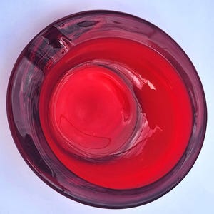 May include: A circular, deep red glass ashtray viewed from directly above. The object features a smooth, translucent surface with a thick, rounded rim and a single wide notch for resting items, showcasing a vibrant crimson hue throughout the glass.