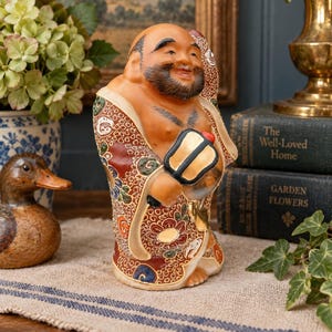 May include: A ceramic figurine of a smiling, bearded figure with a bald head, wearing a robe decorated with intricate red, white, and gold floral patterns. The figure holds a black and gold fan in one hand and a large fish over one shoulder.