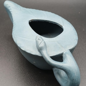 May include: A matte blue ceramic oil lamp with a leaf-shaped body and a curved handle. The top surface features a central opening and embossed leaf vein details. The design includes a small, sculpted snake figure attached to the handle base.