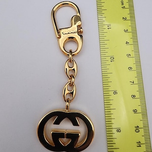 May include: A gold-toned metal keychain featuring the Gucci logo as a pendant, attached to a chain and a lobster clasp. A yellow ruler is placed alongside the keychain, showing the total length is approximately 4.75 inches long.